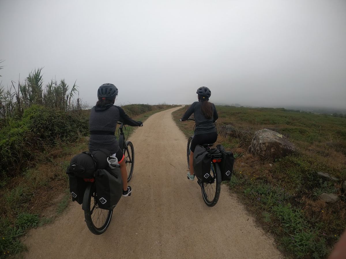 Duas ciclistas num caminho de terra junto à costa, com nevoeiro