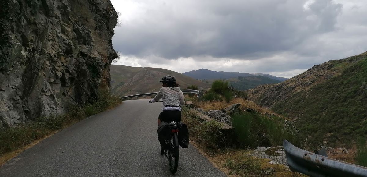 Ciclista numa estrada estreita junto a uma parede de rocha, com céu carregado