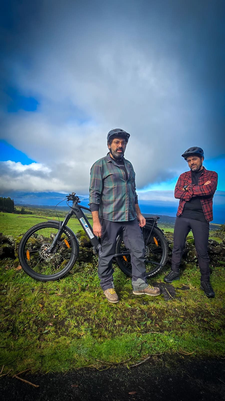 José and Cláudio in helmets next to a BEEQ, with pastures and the sea behind