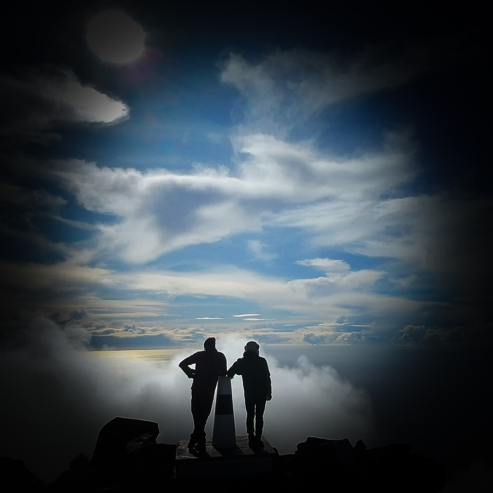 Silhouettes of José and Cláudio by the summit marker, above the clouds
