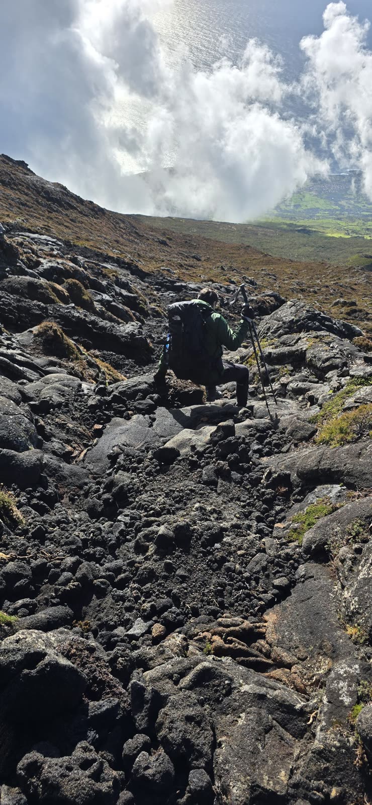 Descending over lava with trekking poles, with clouds and the sea far below